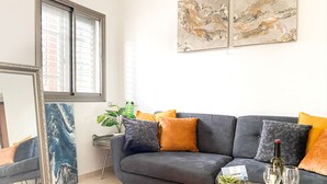 Een woonkamer met een grijze bank, oranje kussens, een glazen salontafel met daop een croissant, wijnglazen en een plant.