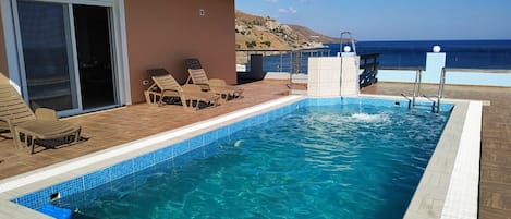 Una piscina rectangular con agua cristalina azul, rodeada por una terraza de madera y sillones de descanso.