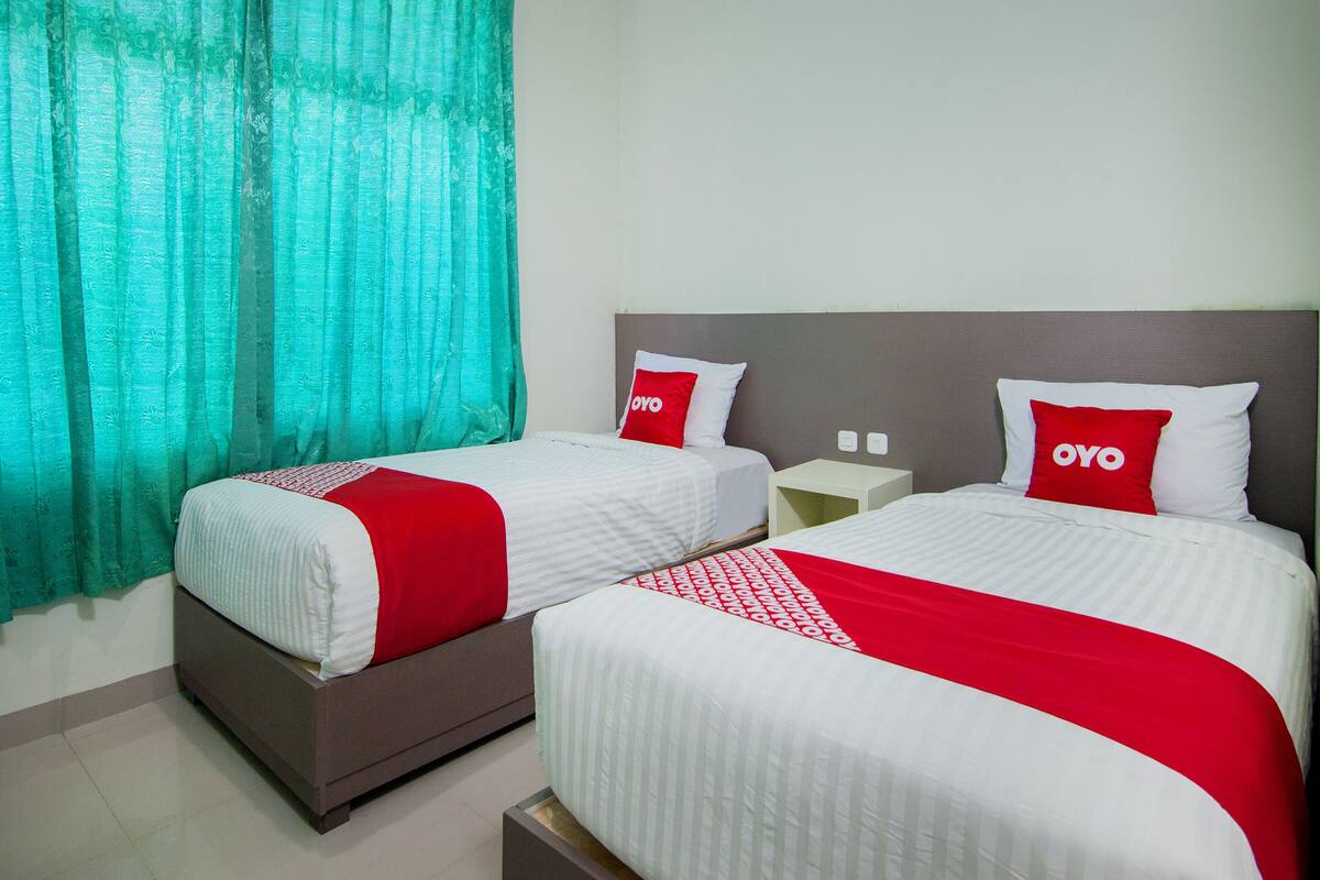 A hotel room with two beds, each with white bedding and red accents, a nightstand, and a window with teal curtains.