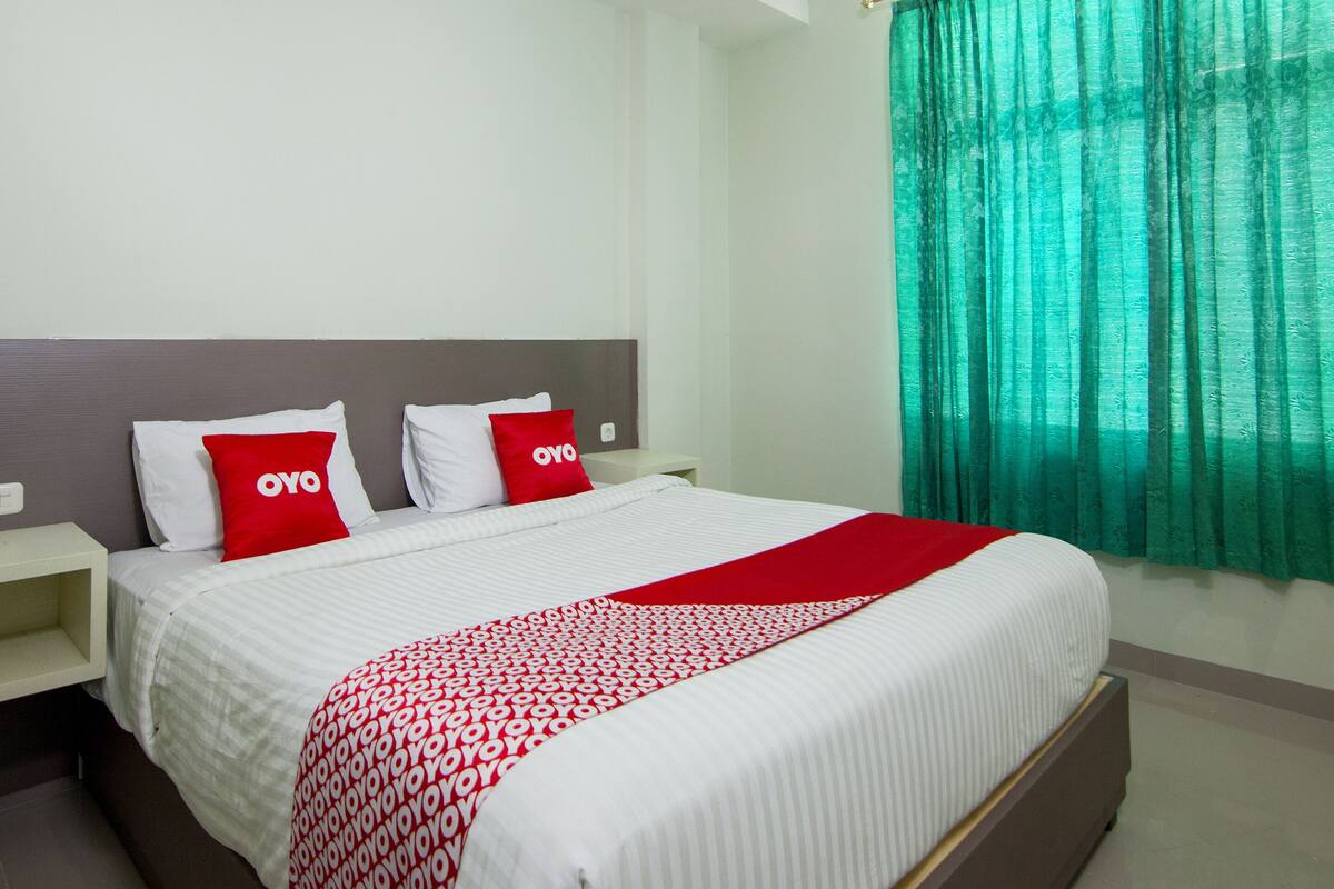 A neatly made bed with white bedding and red and white pillows, a red throw blanket, and a green curtain on the window.