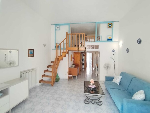 A spacious living room with a blue sofa, a glass coffee table, and a wooden staircase leading to an upper level. - Ischia Ponte Romantic Apartment in the Historical Center and Near the sea (Ischia)