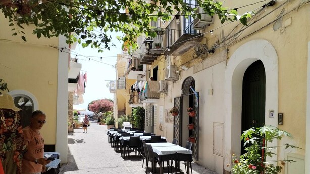 A narrow cobblestone street with outdoor seating, a potted plant, and a building with an arched doorway. - Ischia Ponte Romantic Apartment in the Historical Center and Near the sea (Ischia)