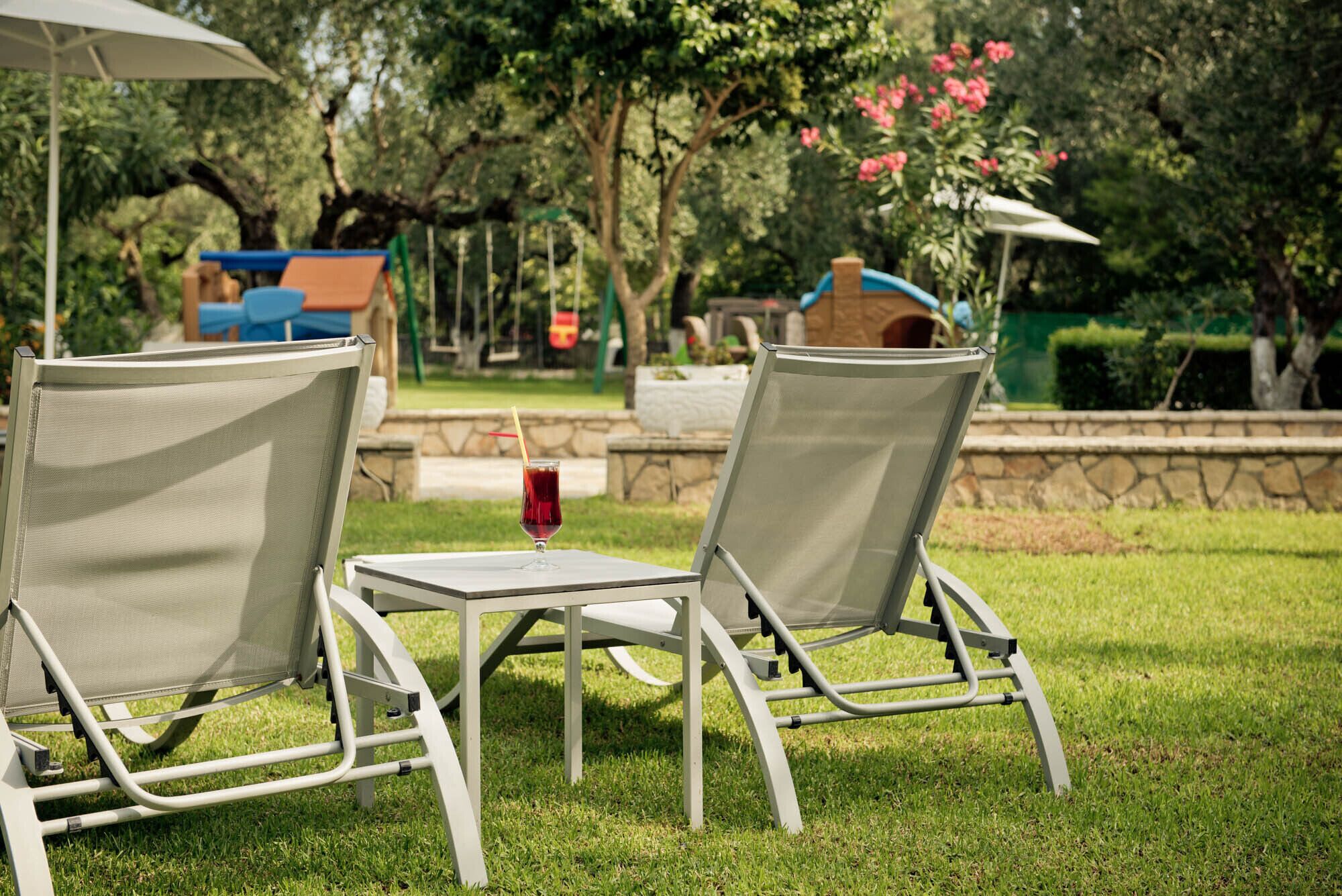 Two outdoor lounge chairs with a table and a drink, set in a grassy area with a playground and stone wall in the background.
