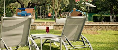 Two outdoor lounge chairs with a table and a drink, set in a grassy area with a playground and stone wall in the background.