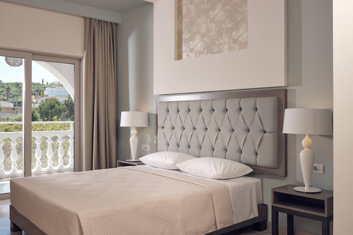 A neatly made bed with a tufted headboard, two bedside lamps, and a view of a balcony with a railing.