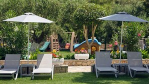 Un espace patio aménagé avec des chaises longues et des parasols, entouré de verdure et agrémenté d’une aire de jeux pour enfants.