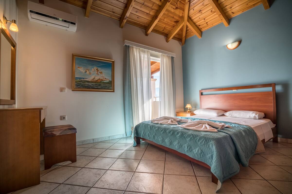 A bedroom with a wooden ceiling, a bed with a blue quilt, a nightstand, a painting, and a window with curtains.