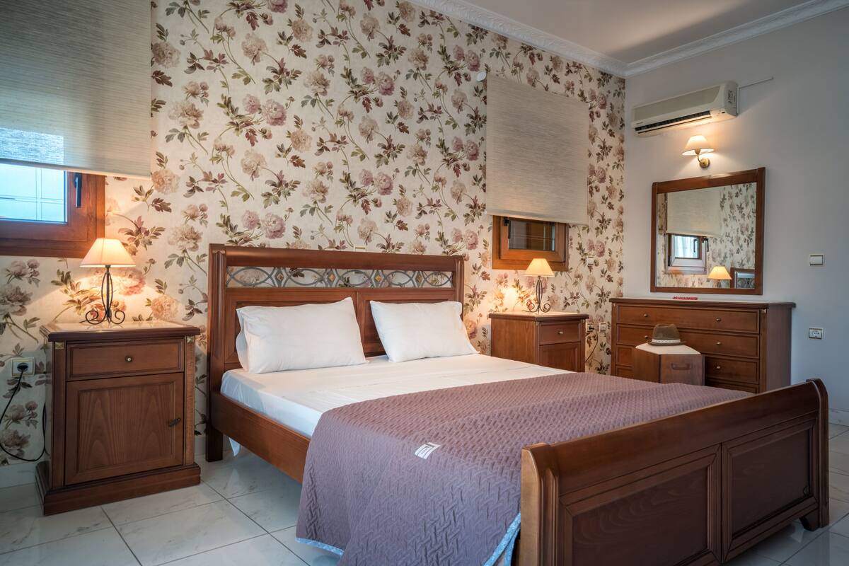A bedroom with a wooden bed, two bedside tables with lamps, a dresser, a mirror, and floral wallpaper.