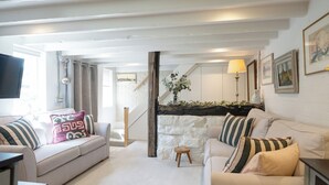 Sala de estar con un sofá blanco, un sofá más pequeño y un taburete de madera. Se ve una escalera que sube a un nivel superior, y un televisor montado en la pared.