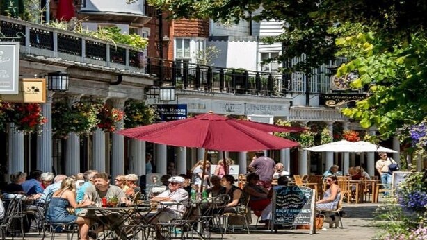 Un café al aire libre con mesas y sillas, rodeado de edificios y vegetación.