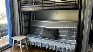 Una cama litera con un edredón a rayas, un taburete de madera pequeño y una ventana con cortinas.