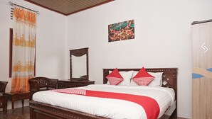 A neatly made bed with a wooden frame and red pillows, a wooden nightstand with a mirror, a wooden wardrobe, and a painting on the wall.
