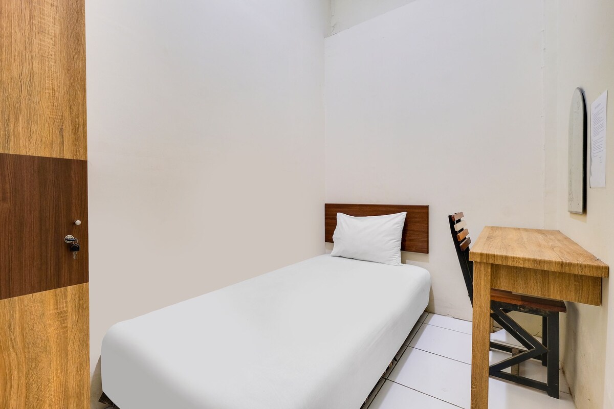 A single bed with a wooden headboard, a small wooden table, and a white wall with a door and a light switch.