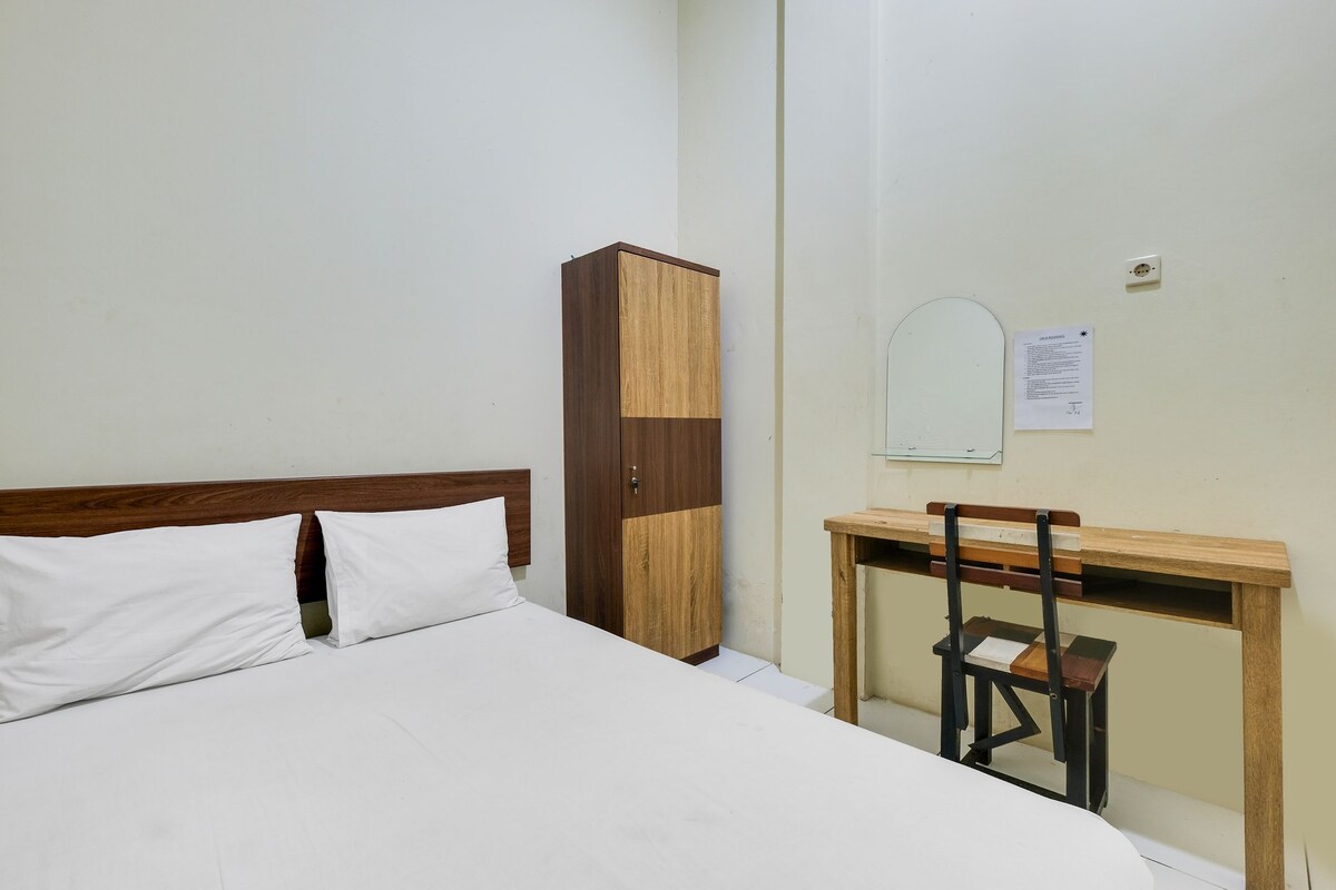 A bedroom with a bed, a wooden wardrobe, a desk with a chair, and a wall-mounted mirror.