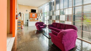 A reception area with a reception desk, two purple sofas, and a large window with blinds. - Hotel O Camel Residence (Semarang)
