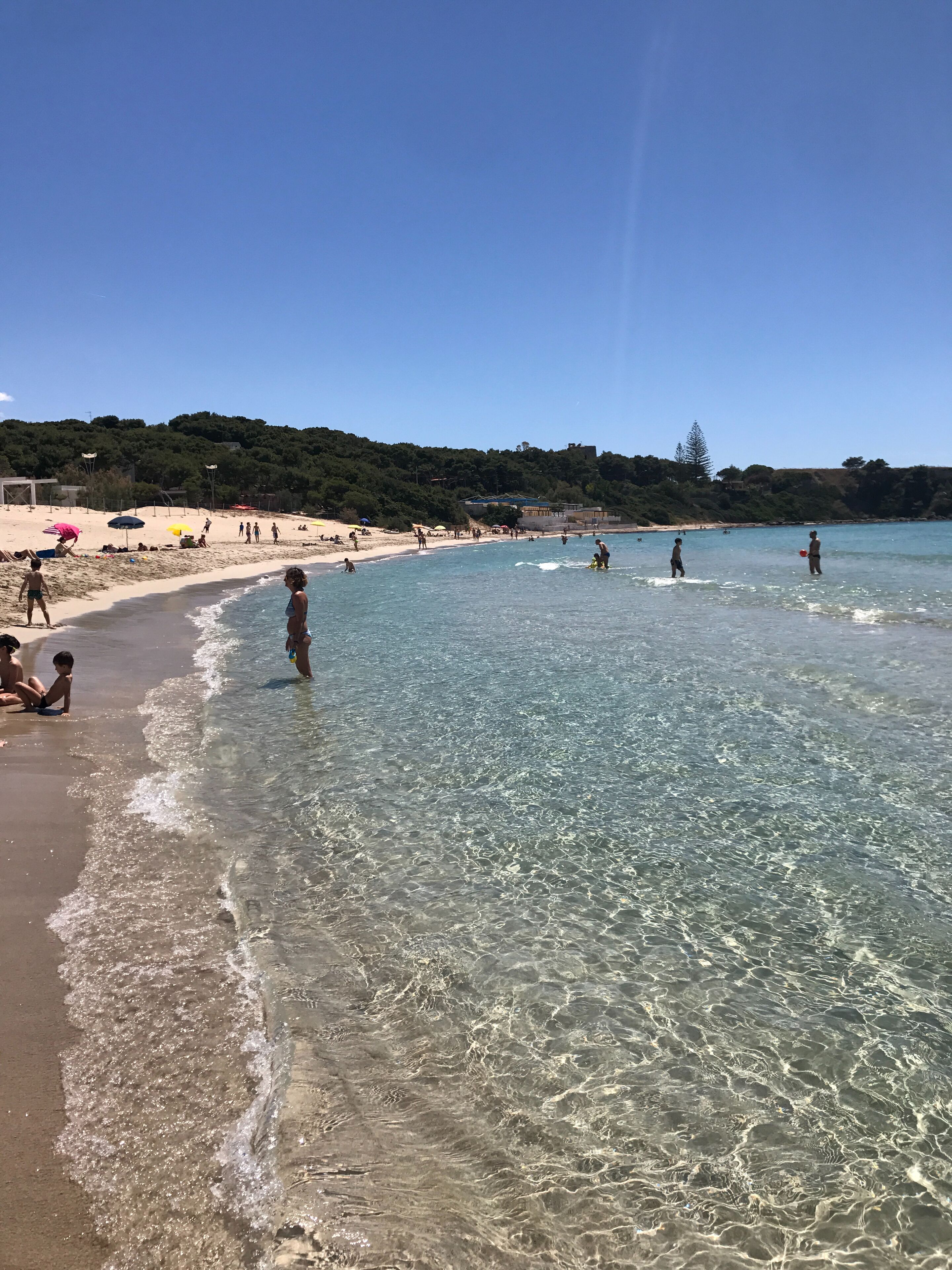 Uma praia com águas cristalinas e rasas, pessoas nadando e caminhando pela orla, e guarda-sóis para banho de sol.