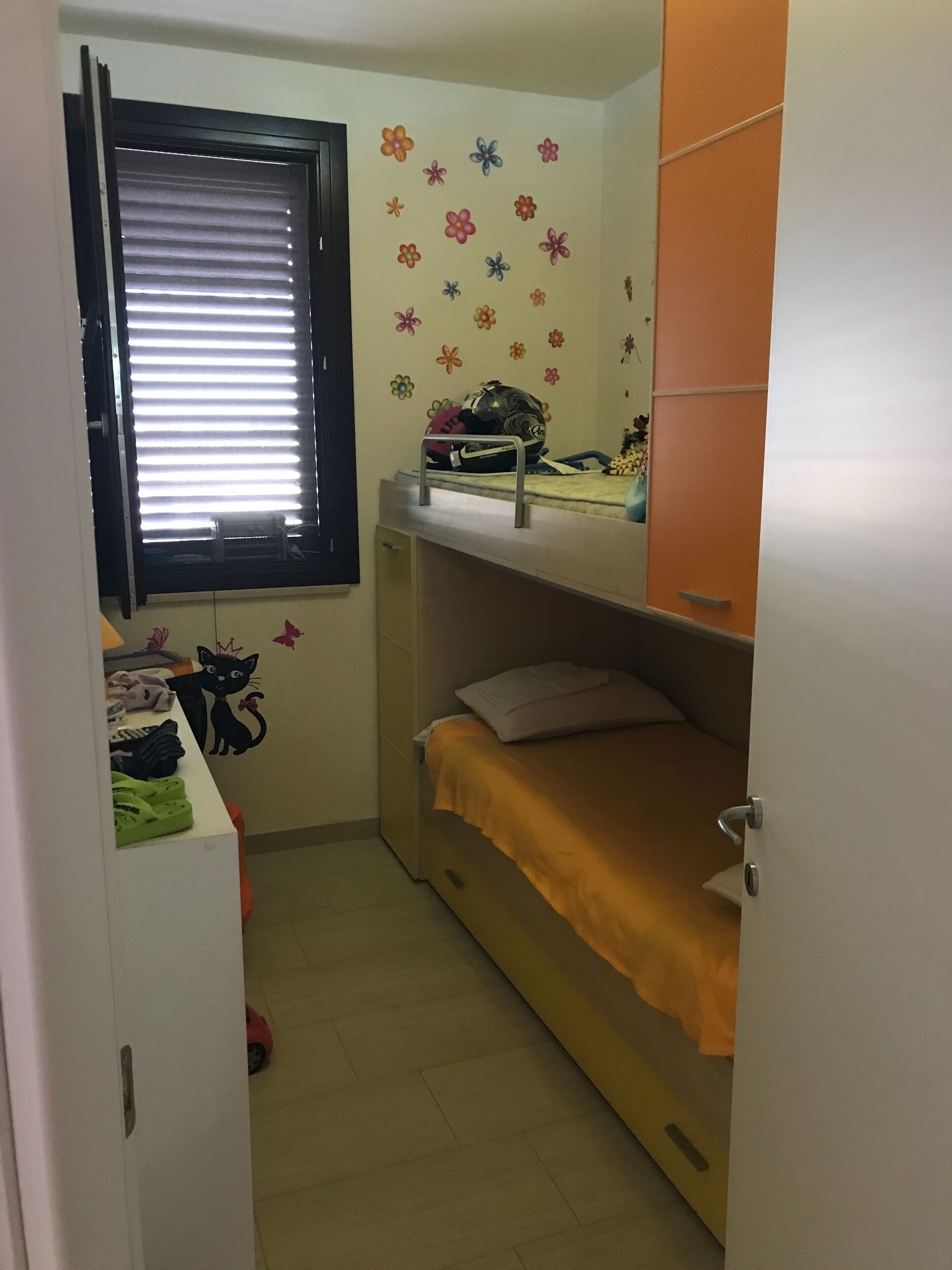 A small, well-lit room with a bunk bed, a window with blinds, a desk with a helmet, and a wall with a cat decal.
