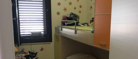 A small, well-lit room with a bunk bed, a window with blinds, a desk with a helmet, and a wall with a cat decal.