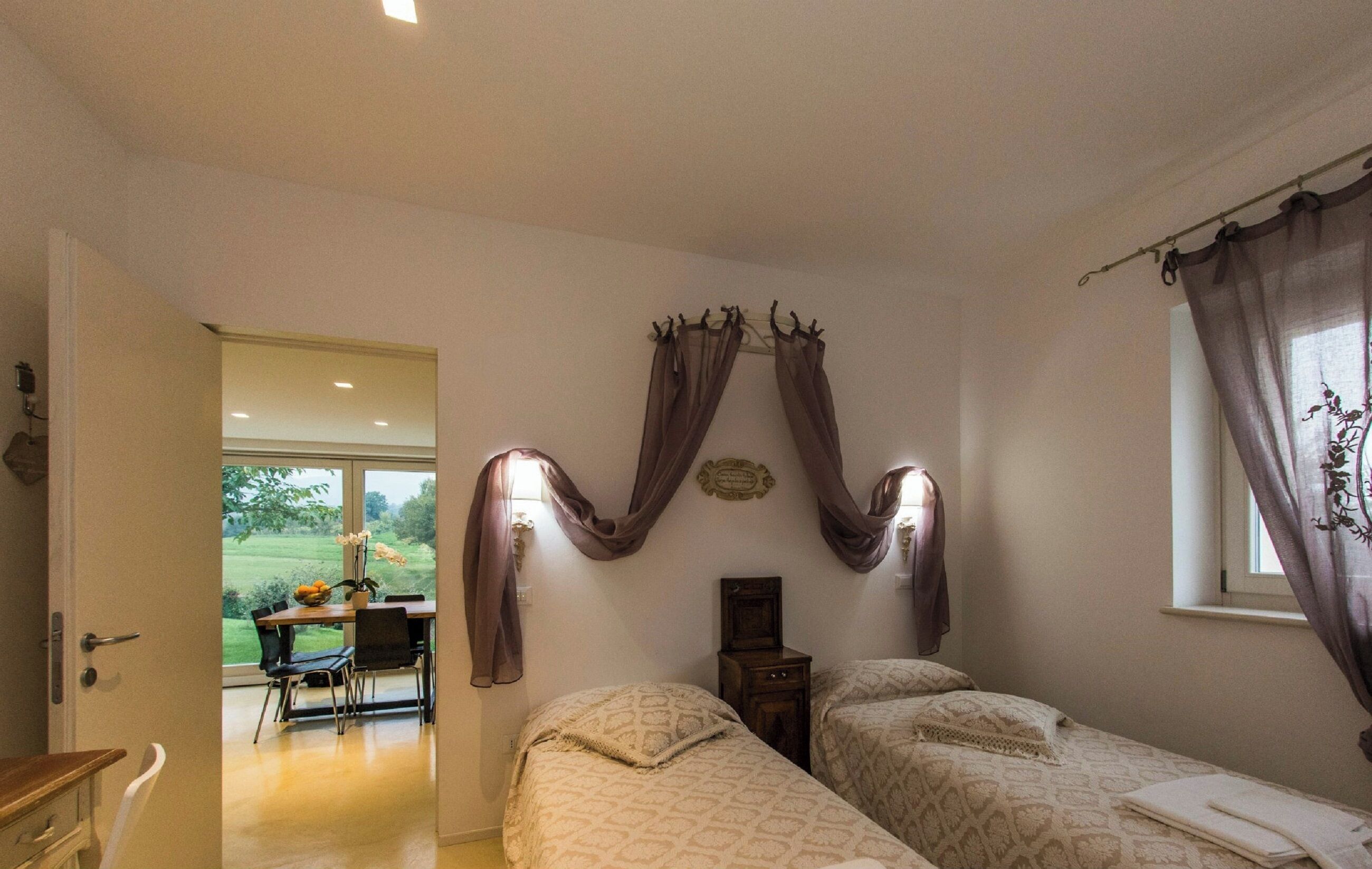 A bedroom with two beds, a window with curtains, a bedside table, and a view of a dining area.