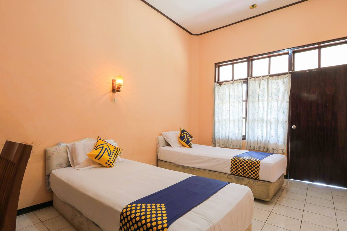 Une chambre d’hôtel avec deux lits simples, une table de chevet, une lampe fixée au mur et une fenêtre avec des rideaux.