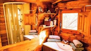 Innenansicht einer Holzhütte mit einem Badezimmer, das ein Waschbecken, einen Handtuchhalter und ein Fenster mit Blick auf Bäume beinhaltet.
