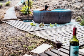 Eine Person entspannt in einem großen, runden Korbsessel in einem Garten mit Holzterrasse, umgeben von kleinen Pflanzen und einer Weinflasche.