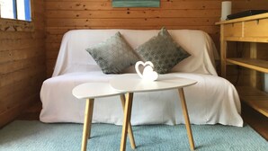 Ein gemütlicher Raum mit einer Holzwand, einem weißen Sofa mit gemusterten Kissen, einem kleinen weißen Tisch mit Herzdeko und einem Bild mit Strandmotiv.