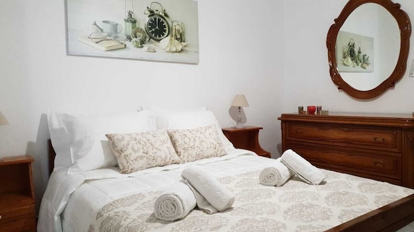 A neatly made bed with rolled towels, a wooden dresser, a mirror, and a painting on the wall.