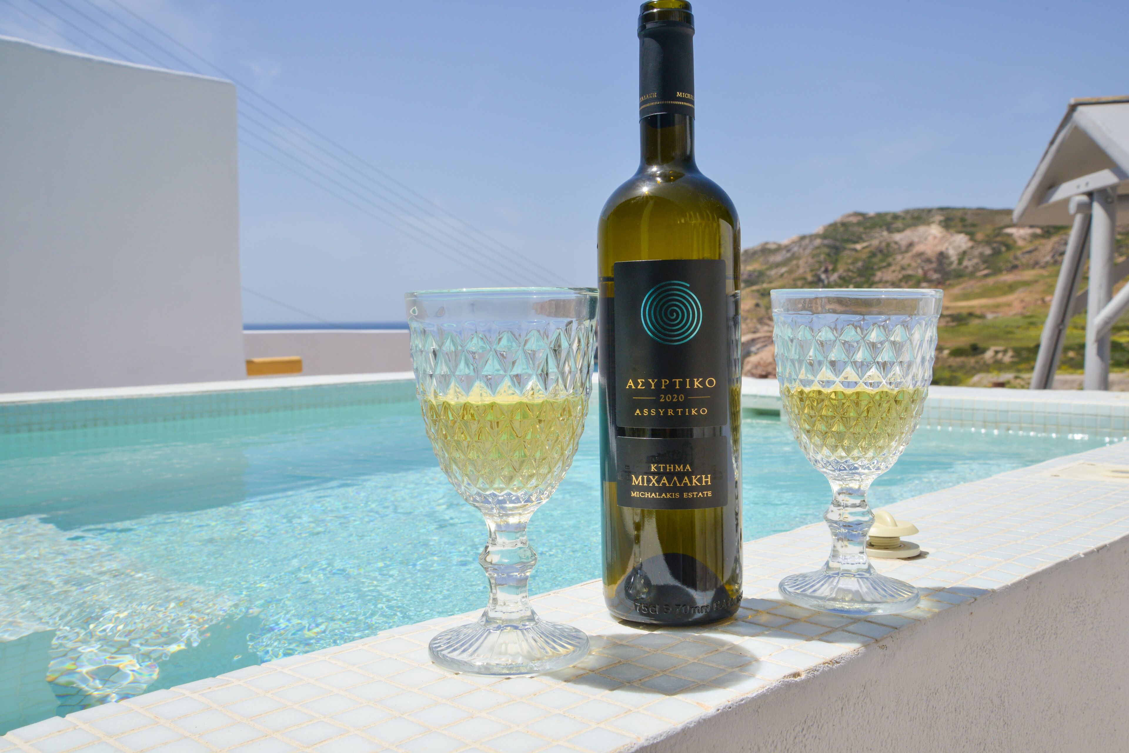 Une bouteille de vin et deux verres posés sur le bord d’une piscine, offrant une vue dégagée sur la mer et les collines en arrière-plan.