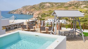 A pool area with clear blue water, lounge chairs, and a view of the sea and hills. - Avgerinos Deluxe Rooms (Milos)