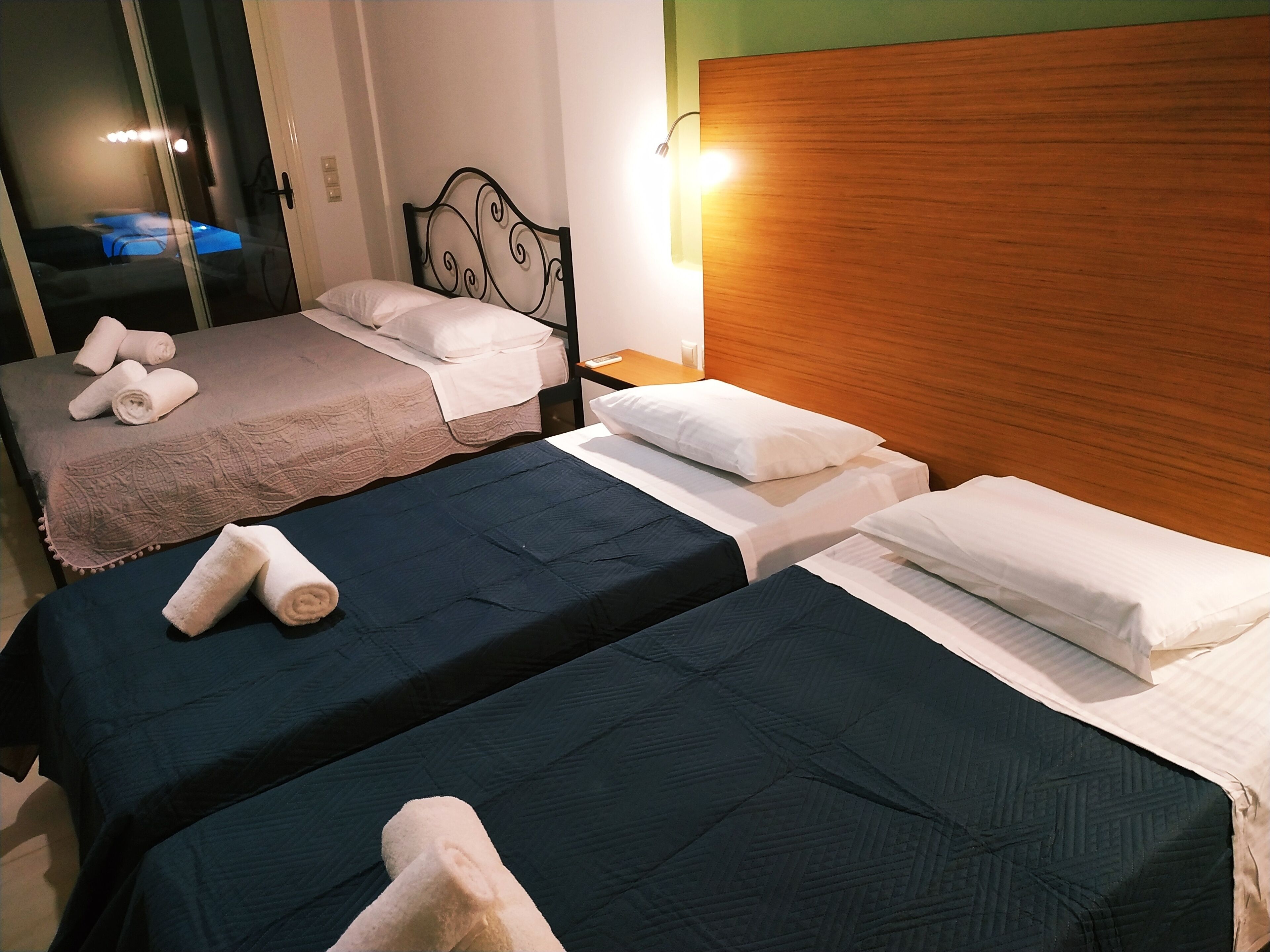 A hotel room with two single beds, each with a dark blue bedspread and white pillows. There is a wooden headboard on one bed and a metal headboard on the other. A wall-mounted light illuminates the room.