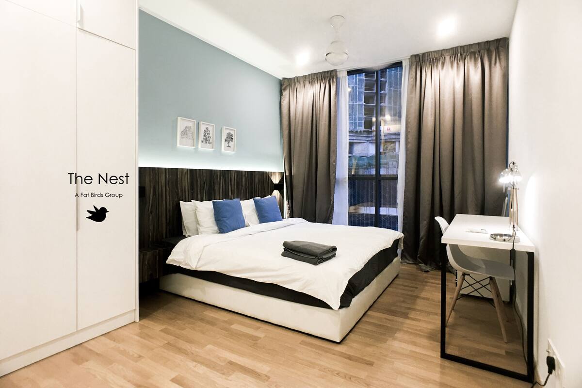 A modern bedroom with a large bed, a desk with a chair, a window with curtains, and a wall-mounted light fixture.