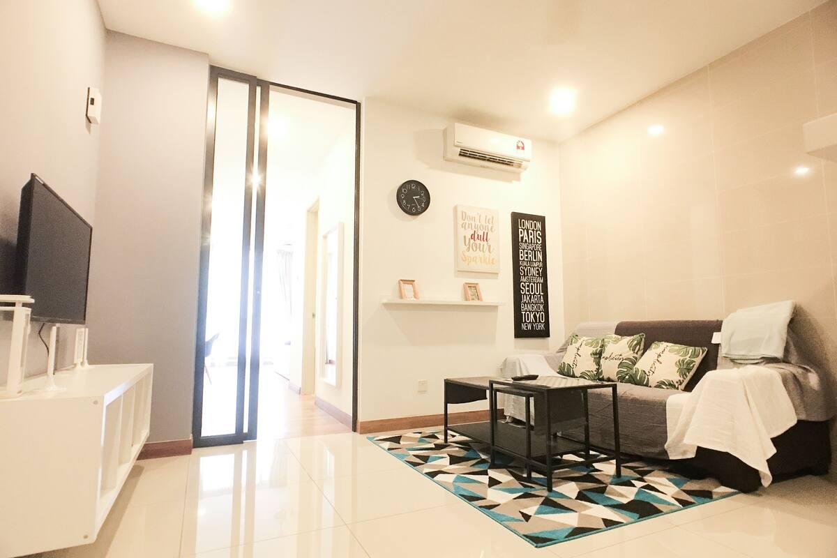 A modern living room with a sofa, a coffee table, and a TV on a stand. There is a patterned rug on the floor and a wall-mounted air conditioner.