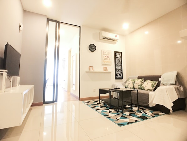 A modern living room with a sofa, a coffee table, and a TV on a stand. There is a patterned rug on the floor and a wall-mounted air conditioner.