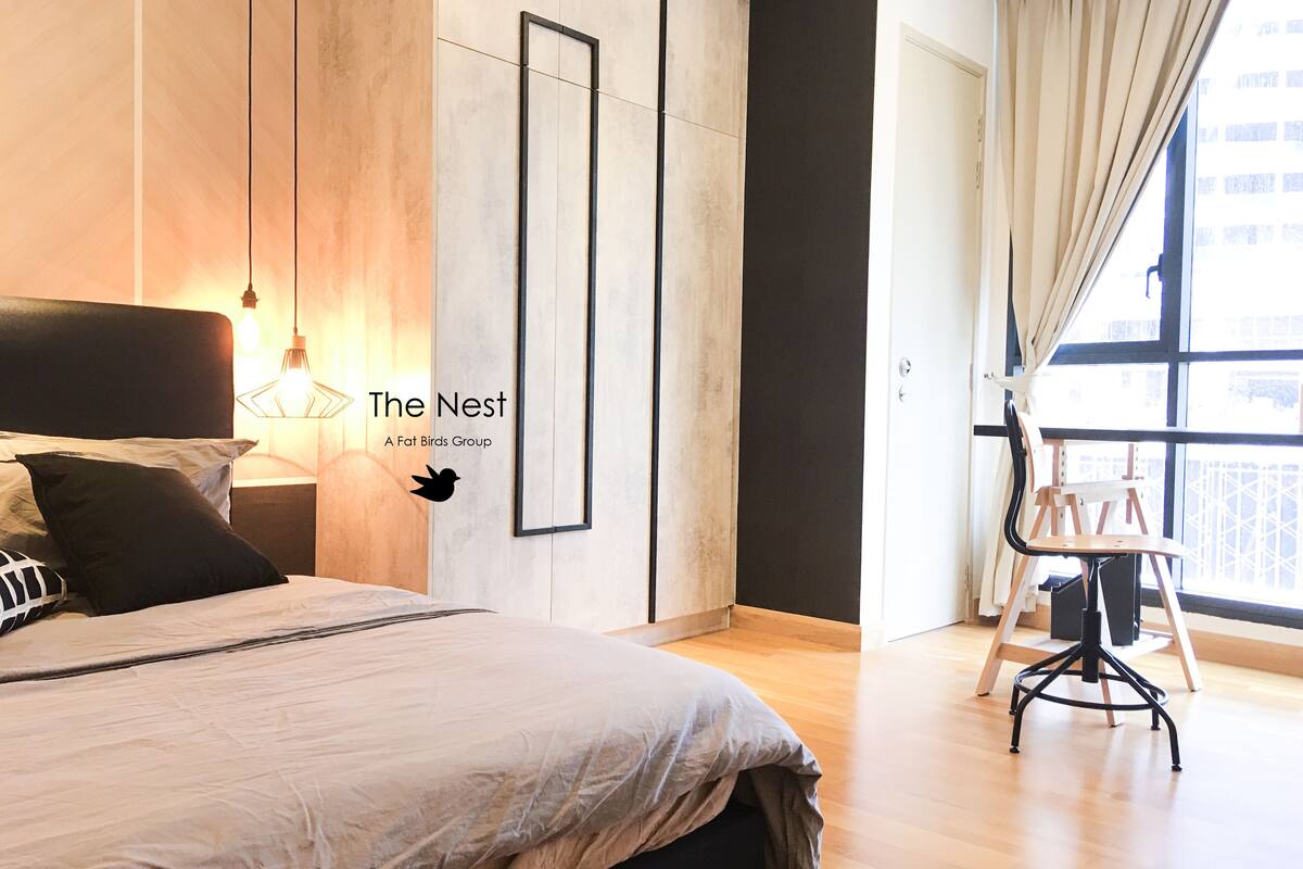 A modern bedroom with a large bed, a desk, and a chair. There is a framed picture on the wall and a window with curtains.