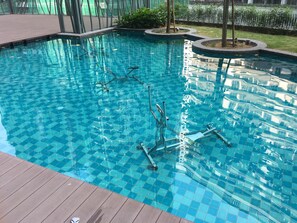 A swimming pool with clear blue tiles, surrounded by a wooden deck and a grassy area with trees.