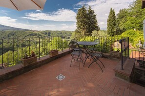 Terraço com mesa e cadeiras, rodeado por uma grade de metal, com vista para uma paisagem verdejante.