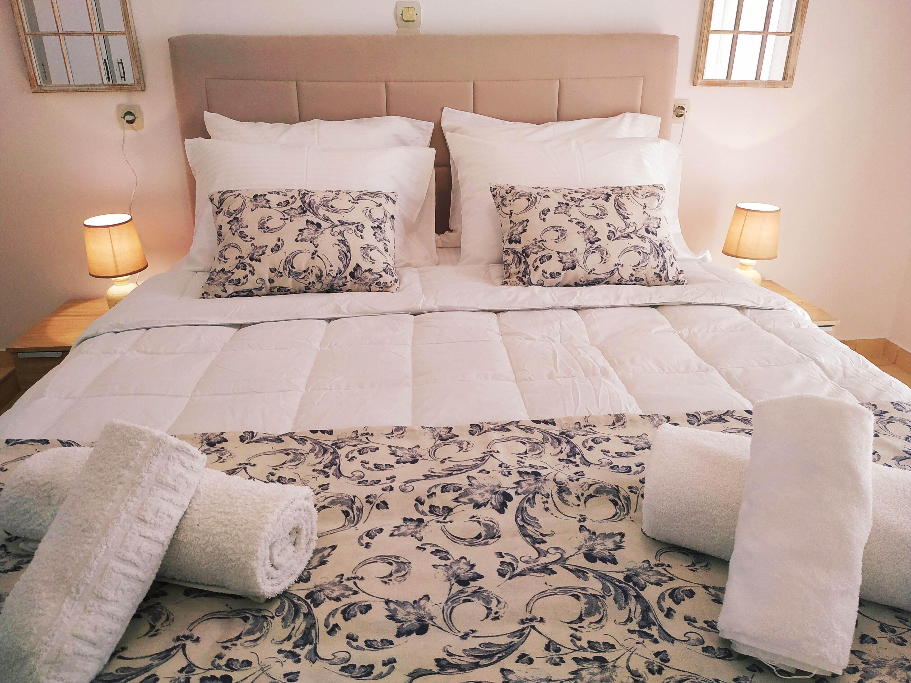 A neatly made bed with a patterned duvet, two pillows with a similar design, and two rolled towels on the bed.