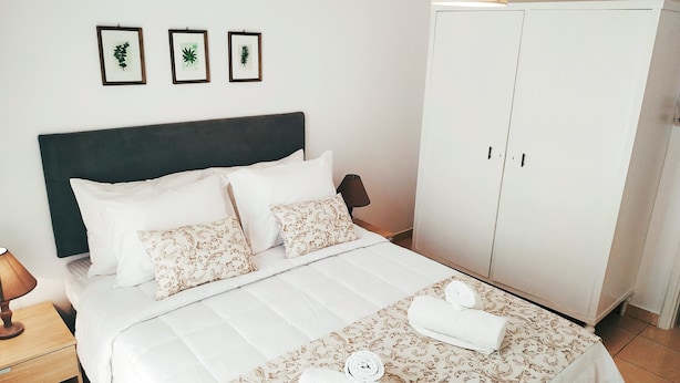 Una cama bien hecha con ropa blanca y almohadas estampadas, una lámpara de noche y un armario con espejo.