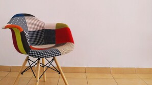 Una silla colorida con diseño patchwork en una habitación con suelo de baldosas y un cuadro grande y vibrante que representa una escena imaginativa con animales, globos aerostáticos y un barco.