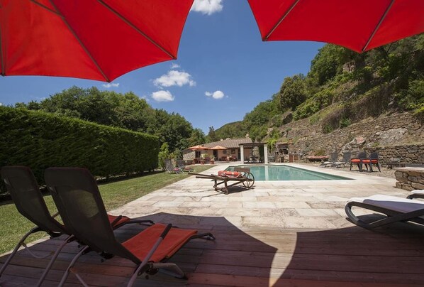 Ein Poolbereich mit Liegestühlen und Sonnenschirmen, umgeben von einer Stegterrasse und Blick auf einen Hügel.