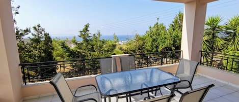 A balcony with a glass table and chairs, offering a view of the sea and greenery.