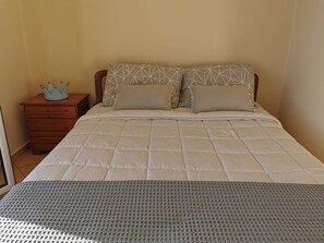 A neatly made bed with a patterned headboard, a wooden nightstand with drawers, and a decorative crown on the nightstand. - Amazing House Erato On The Sea South Creta (Archanes-Asterousia)