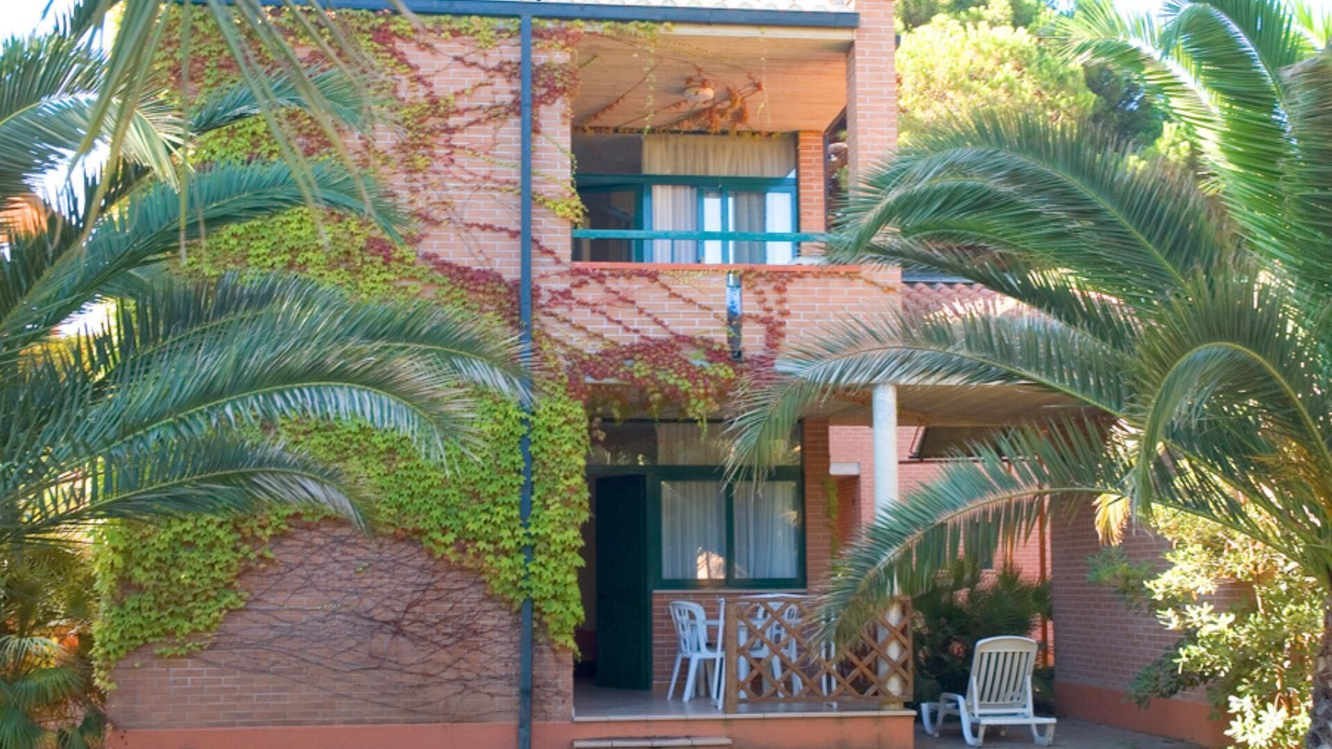 Edificio de dos pisos con fachada de ladrillo cubierta parcialmente de hiedra. Cuenta con un balcón con mesa y sillas, y un pequeño patio con una silla y una mesa.