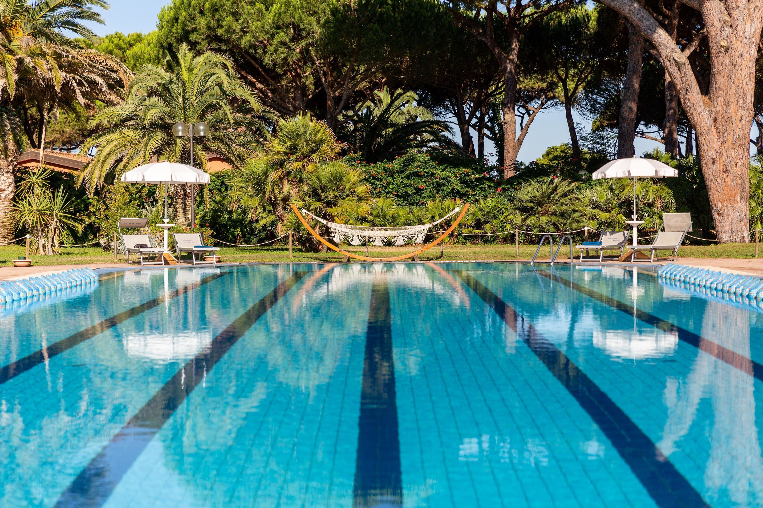 Piscina con sillas de descanso y sombrillas, rodeada de árboles y follaje.