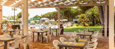 An outdoor restaurant with tables and chairs, a pool in the background, and greenery.