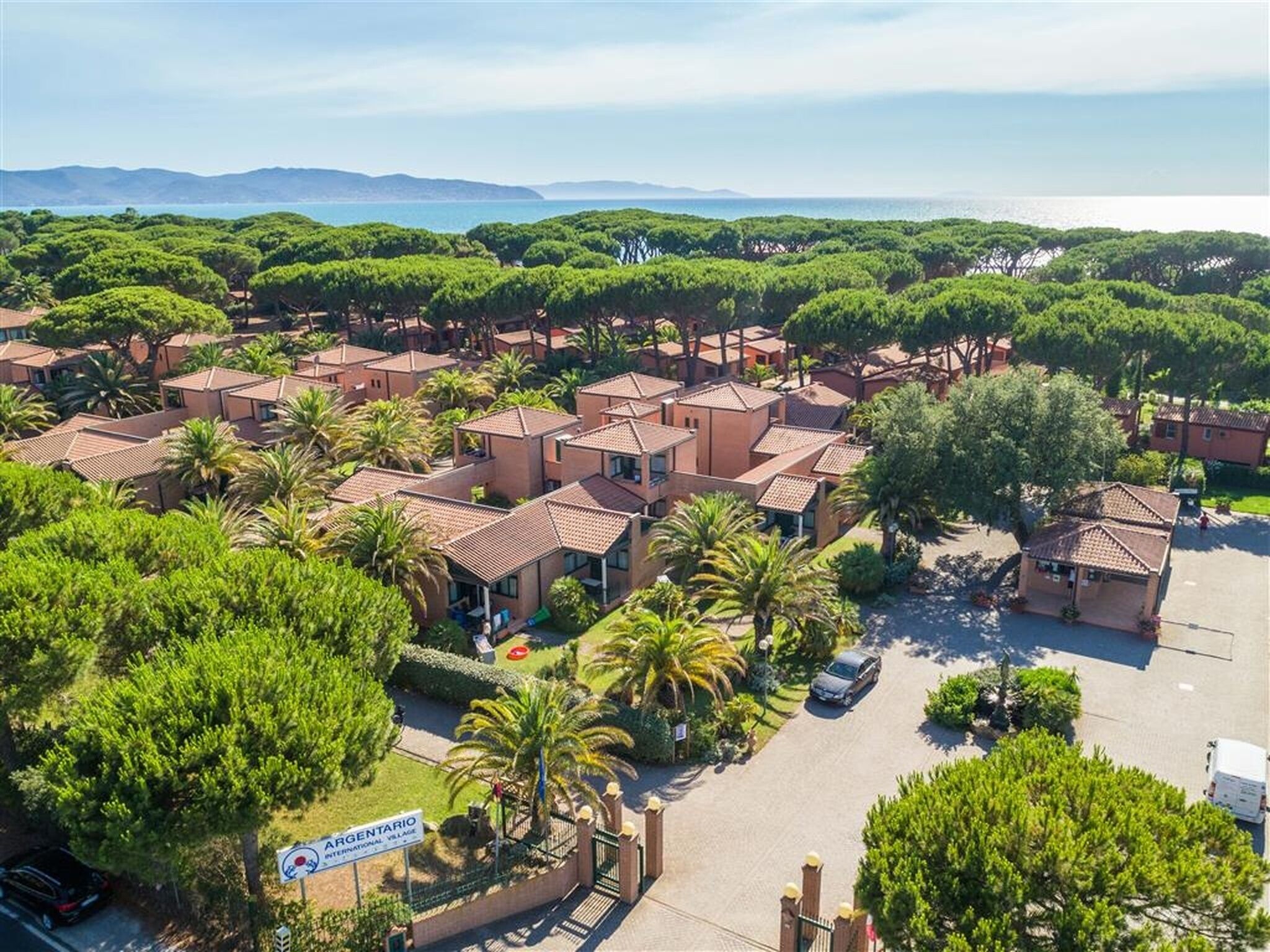 A residential area with multiple houses, a parking area, and a sign indicating 'ARGENTARIO INTERNATIONAL VILLAGE.'