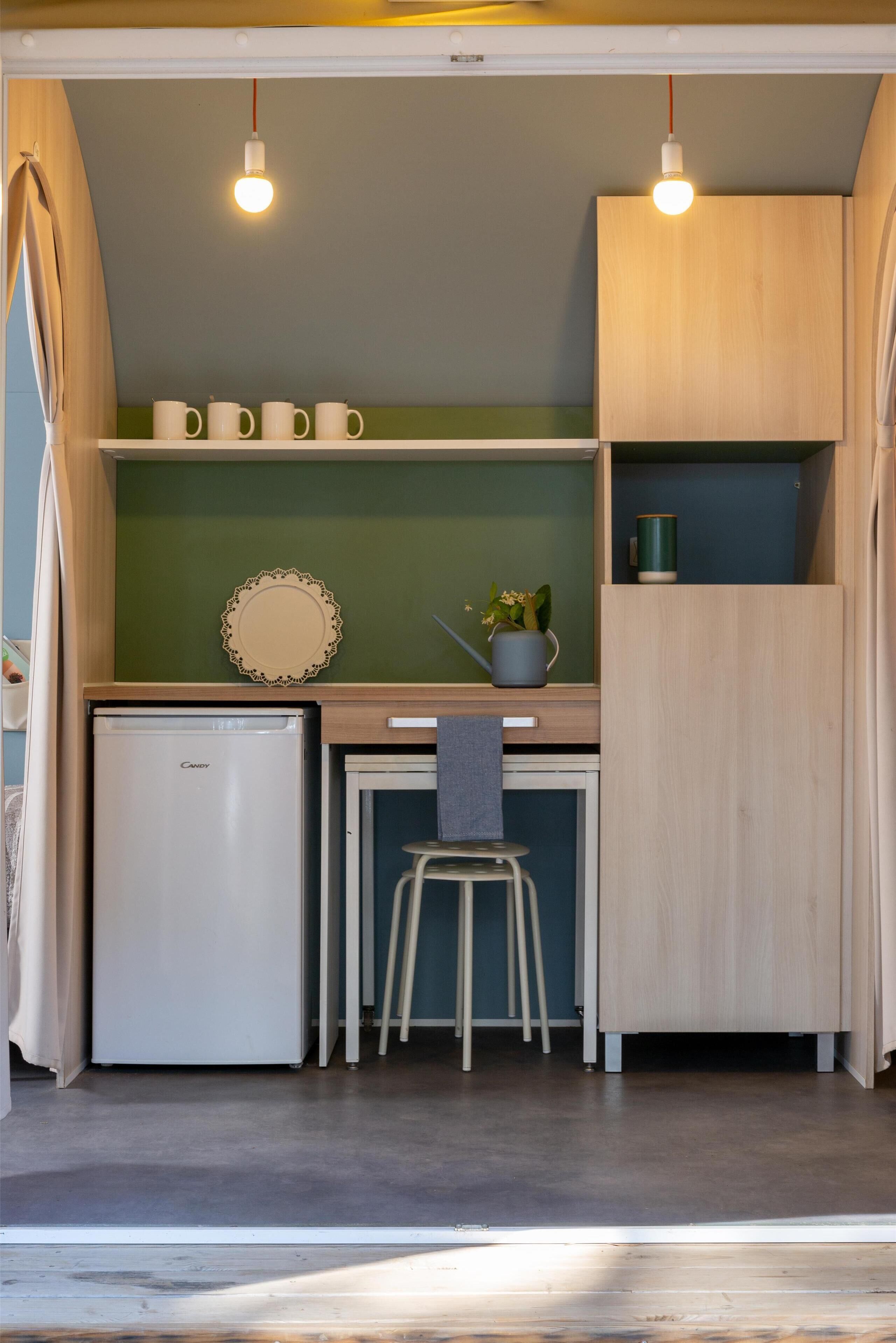 Una kitchenette compacta con refrigerador, un taburete pequeño, una planta en maceta y una lámpara.