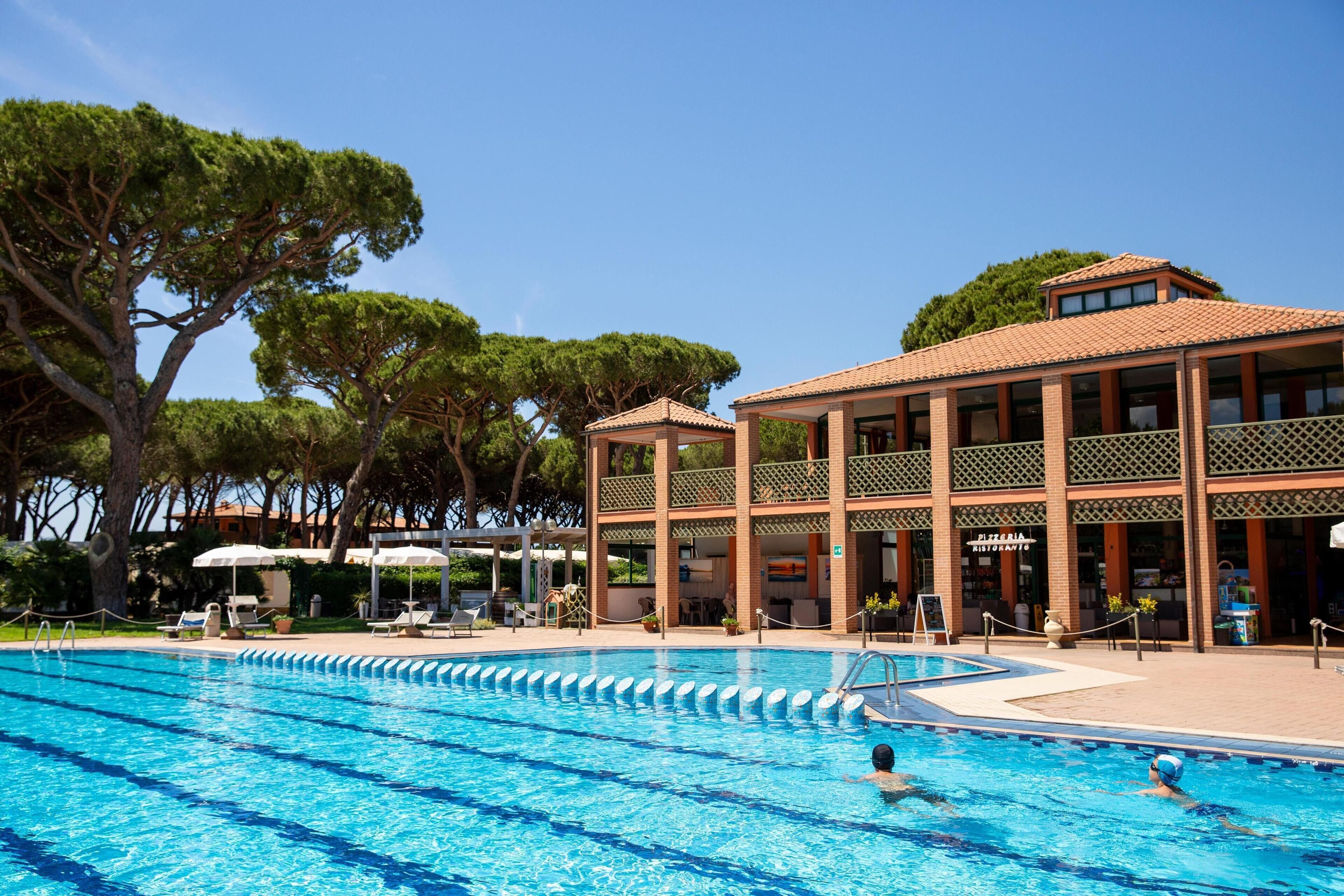 Ein Swimmingpool mit klarem, blauem Wasser, umgeben von Liegestühlen und Sonnenschirmen. Im Hintergrund ist ein Gebäude mit einem Restaurantschild zu sehen.