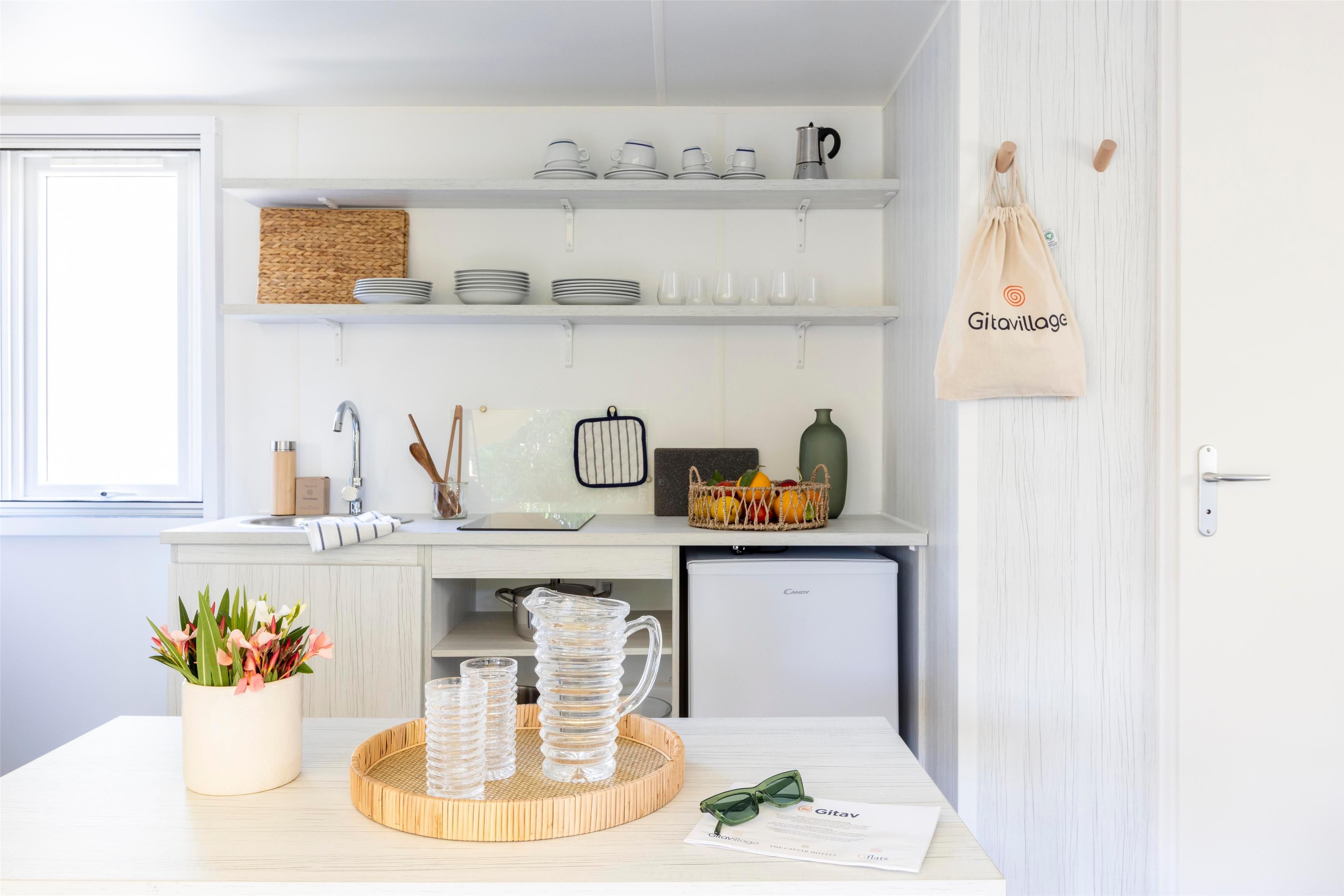 Une cuisine moderne avec des meubles blancs, un évier et un plan de travail sur lequel se trouvent une carafe en verre, des verres et un panier de fruits.